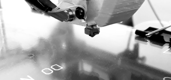 Fotografia em preto e branco de um cabeçote de uma impressora 3D como dispositivo extrusor, com o bico (centro) acoplado ao extrusor (em cima).